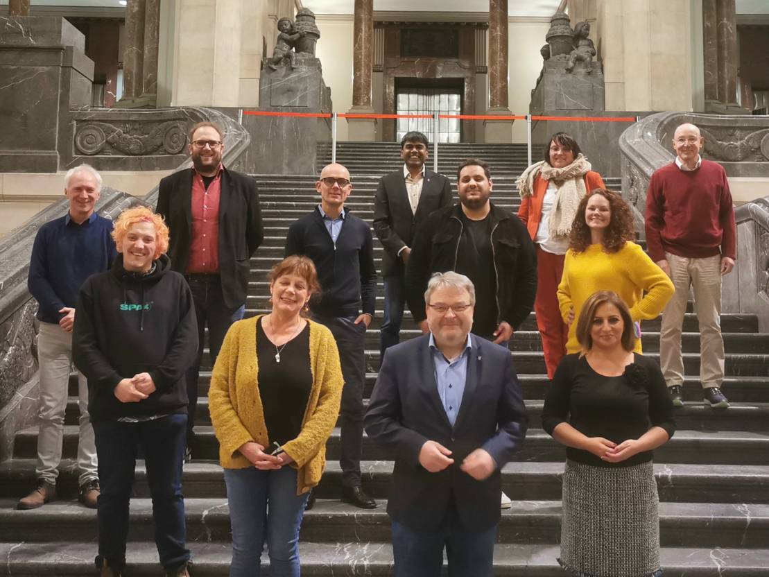 Zwölf Menschen, acht Männer und vier Frauen, stehen in einigem Abstand auf einer großen Freitreppe. Sie posieren für ein Gruppenbild und schauen in die Kamera.